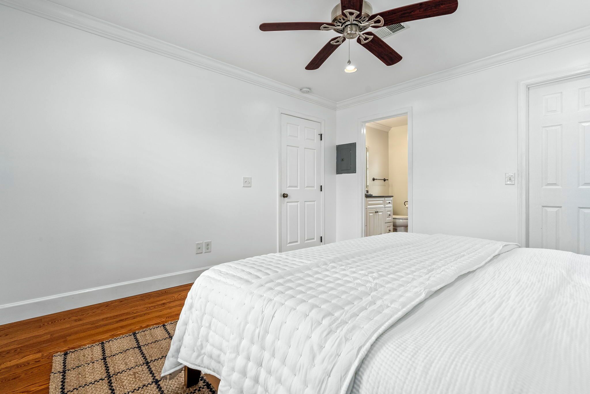 A bedroom with a bed, wooden flooring, white walls, a ceiling fan, and a door leading to a bathroom.