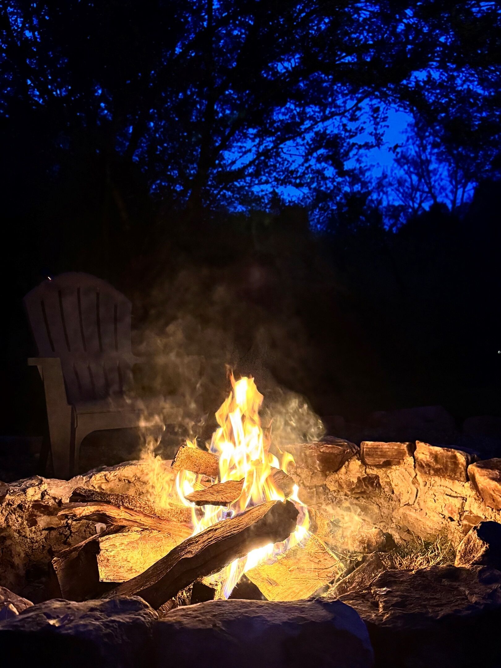 A campfire with logs and rocks, a plastic chair, and a dark sky with trees in the background.