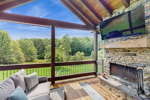A covered deck with a stone fireplace, a TV mounted on the wall, and a view of a green landscape.