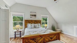 Un dormitorio con cama de madera, dos ventanas con persianas, una mesita de noche con lámpara y vistas a la vegetación.