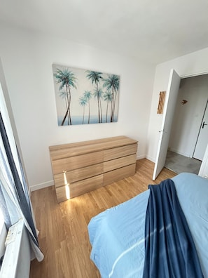 Une chambre avec une commode en bois, un lit recouvert dâune couverture bleue et un tableau reprĂ©sentant des palmiers accrochĂ© au mur.