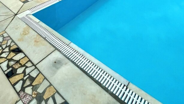 En pool med fri utsikt över havet, en byggnad i sten med ett fönster och en uteplats med kakelplattor.