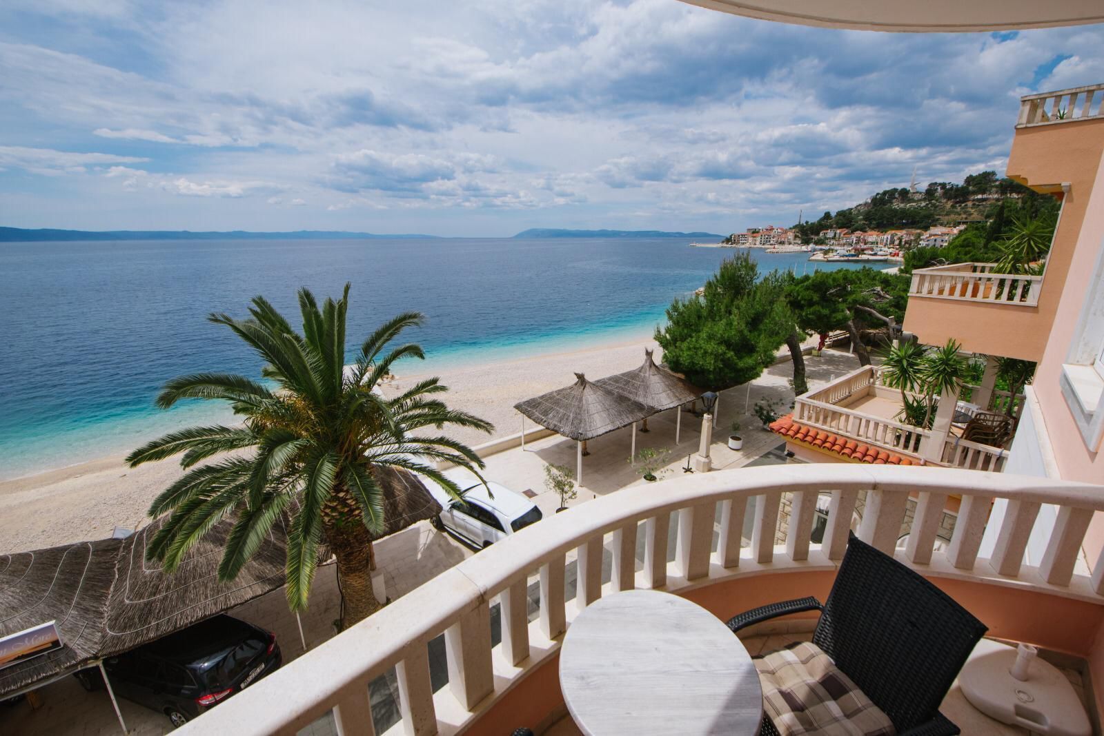 A balcony with a view of a beach and the sea, a palm tree, and a table with a chair.