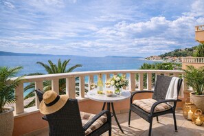 Ein Balkon mit Korbstühlen, einem Tisch mit Flasche und Blumen sowie Blick auf das Meer und sanfte Hügel in der Ferne.
