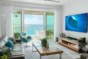 A modern living room with a leather sofa, a glass coffee table, and a large TV screen displaying an underwater scene. - Beachcomber 28 by Grand Cayman Villas (Seven Mile Beach)