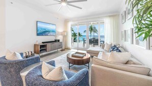 A modern living room with a ceiling fan, a flat-screen TV, and a view of the ocean through large windows. - Beachcomber 7 by Grand Cayman Villas (Seven Mile Beach)