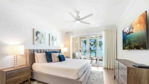 A modern hotel room with a large bed, a ceiling fan, a desk, and a view of the outdoors. - Beachcomber 7 by Grand Cayman Villas (Seven Mile Beach)