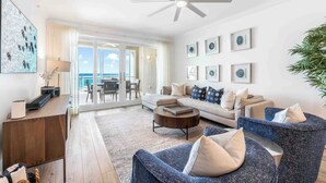 A spacious living room with a large sofa, a coffee table, and a TV on a wooden stand. There is a balcony with a view of the ocean. - Beachcomber 7 by Grand Cayman Villas (Seven Mile Beach)