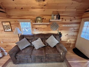 A cozy cabin interior with a brown leather sofa, a woven rug, wooden walls, a window, and a shelf with decorative items.