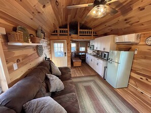 Ein gemütliches Blockhaus-Interieur mit einer braunen Couch, Holzwänden, einer Küche mit weißen Schränken und einem Deckenventilator.
