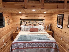 A wooden-paneled room with a bed, nightstands, a wall-mounted TV, and a framed picture.