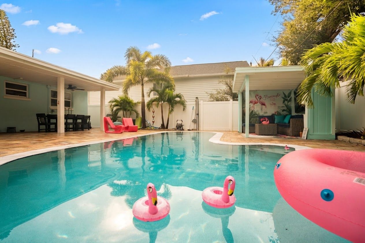 A swimming pool with two pink flamingo floats, surrounded by palm trees and a covered patio area.