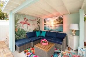 Ein überdachter Außenbereich mit einer blauen Couch, einem Couchtisch und einer Wandmalerei mit Flamingos und Sonnenuntergang.