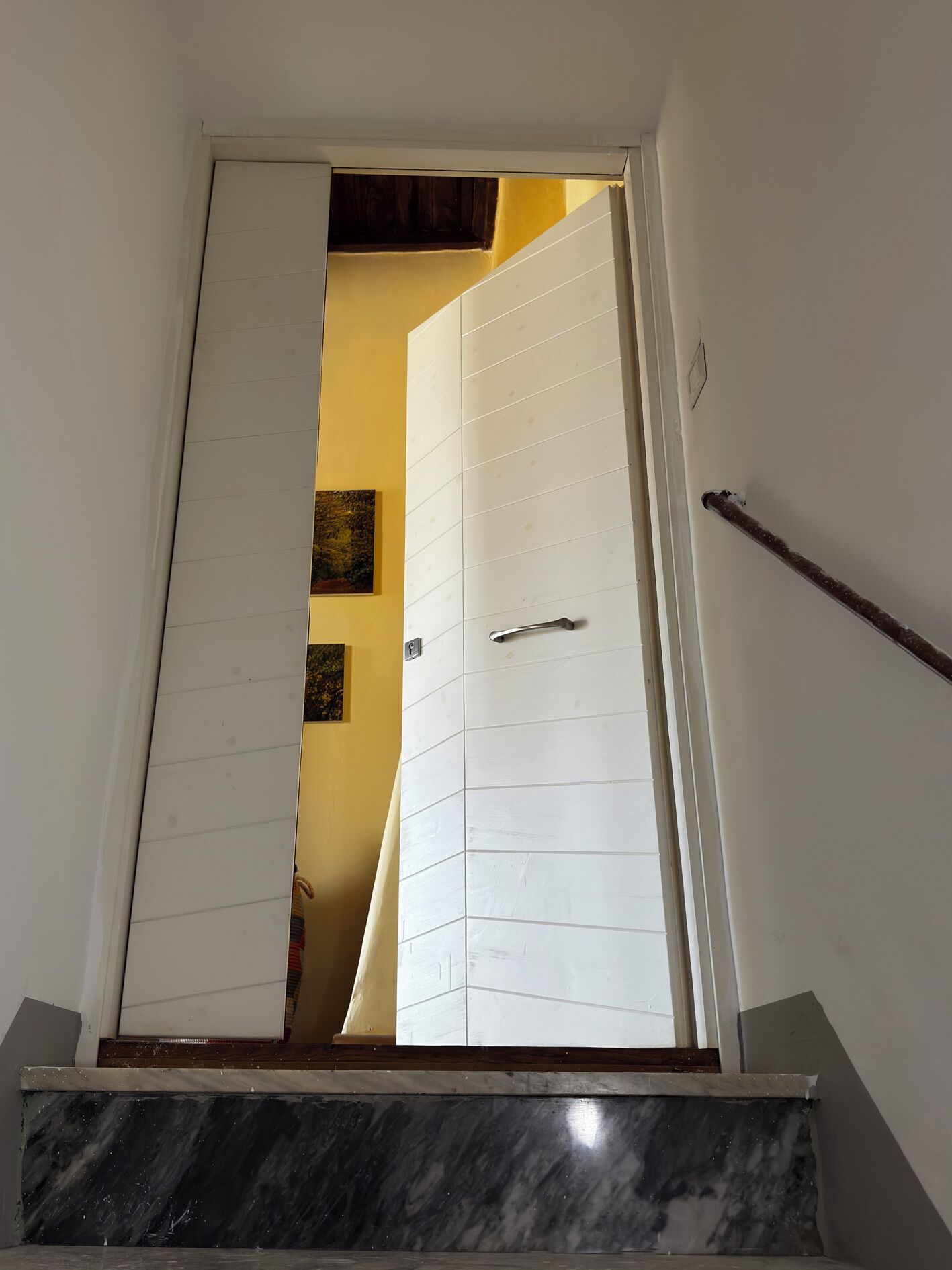 Un escalier menant à une porte blanche située en haut, qui donne sur une pièce aux murs jaunes et ornée de deux tableaux encadrés.