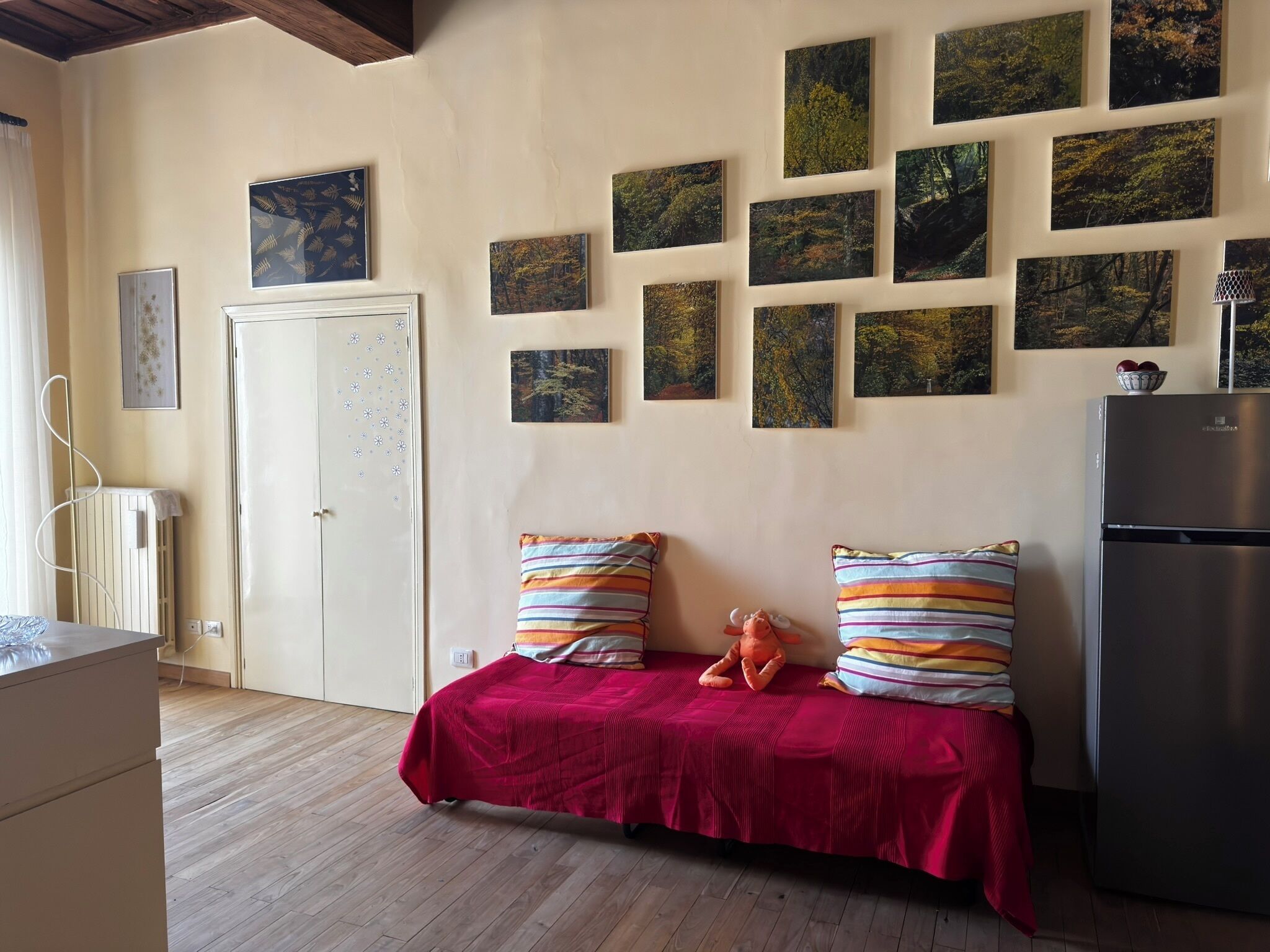 Una stanza con pavimento in legno, un divano con cuscini a righe, un frigorifero e una parete decorata con diverse cornici.