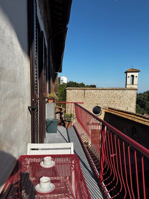 Ein Balkon mit roter Geländer, ein Tisch mit Tassen und Blick auf ein Gebäude mit einem Glockenturm.