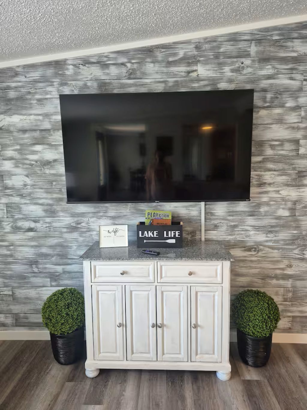 Un salon avec un téléviseur à écran plat fixé à un mur texturé, un meuble blanc avec des tiroirs, deux plantes en pot et un petit panneau sur le meuble.