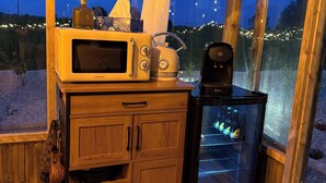 Ein Holzkabinett mit einer Mikrowelle, einem Toaster und einer Kaffeemaschine, ein Kühlschrank mit Flaschen sowie ein Fenster mit Lichterketten.
