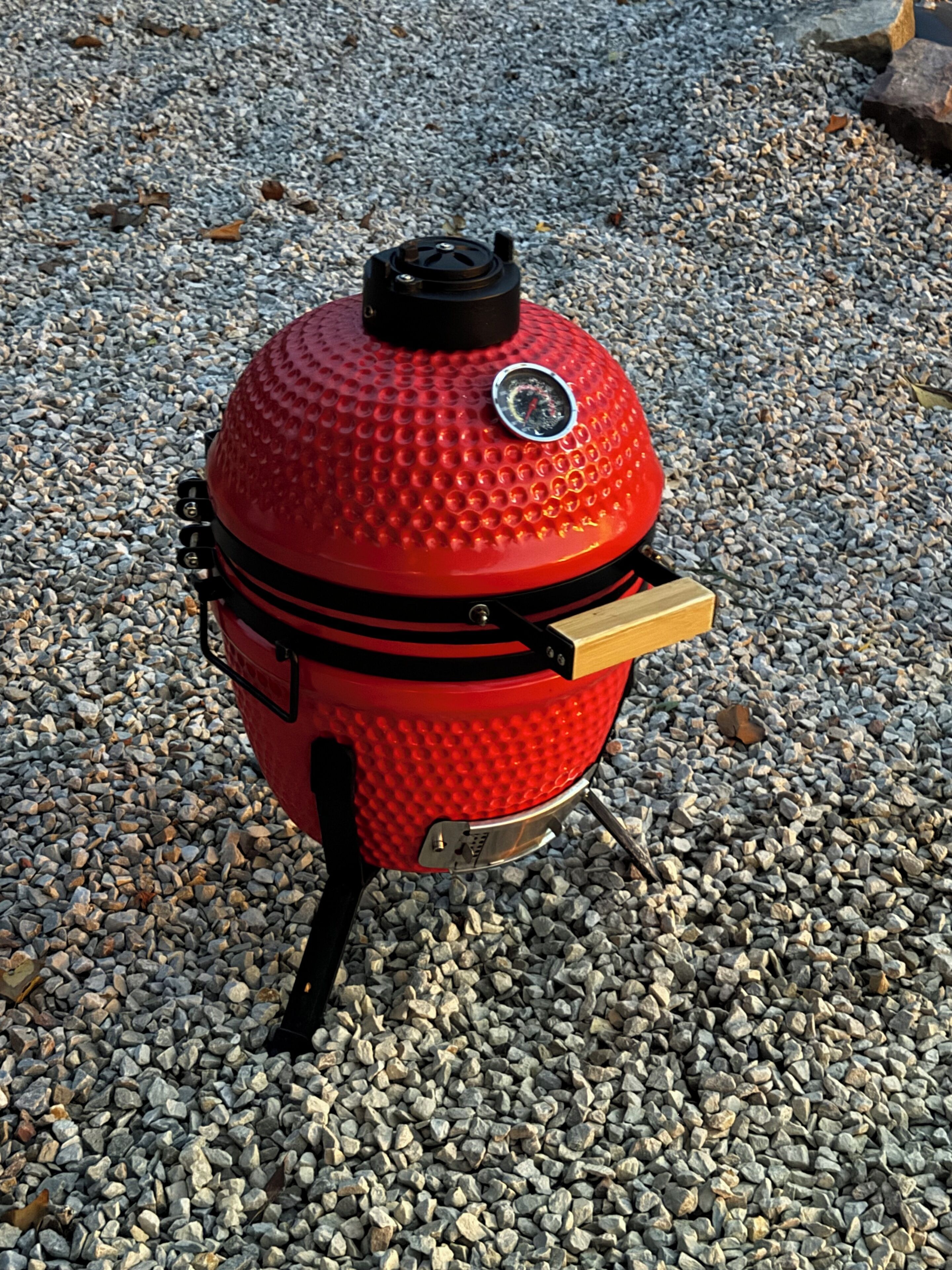 A red barbecue grill with a thermometer and a handle, placed on a gravel surface.