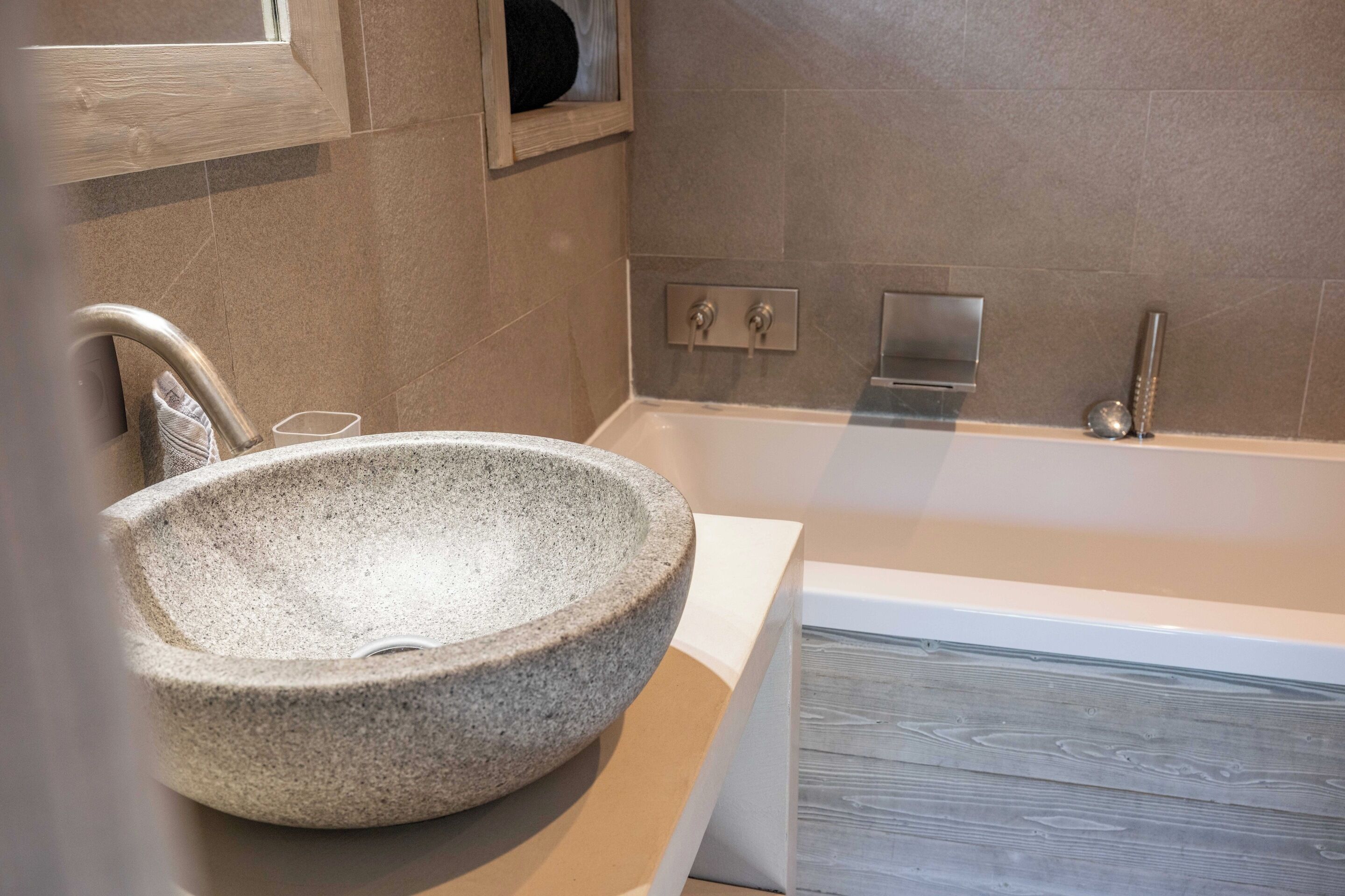 A stone basin sink with a modern faucet, a bathtub with a wooden floor, and a wall-mounted control panel.