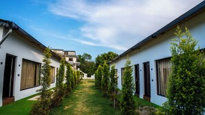 A courtyard with two-story buildings, a clear sky, and a pathway lined with trees. - Corbett Retreat (Kotabagh)
