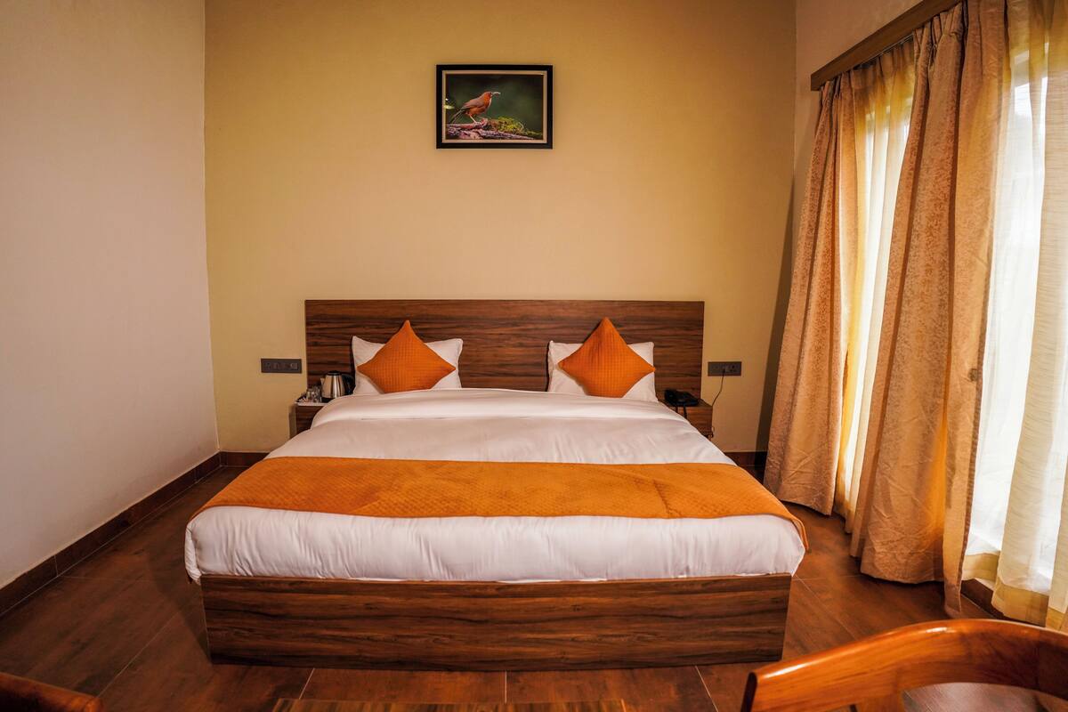 A hotel room with a wooden bed, white bedding, orange pillows, a nightstand, a framed picture on the wall, and curtains on the window.