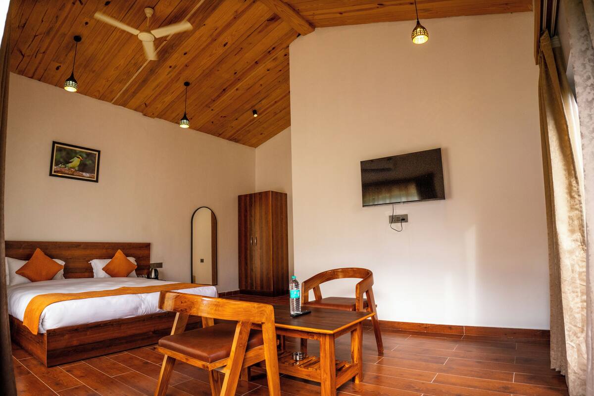 A bedroom with a wooden ceiling, a bed with white bedding, a wooden table with a chair, and a TV mounted on the wall.