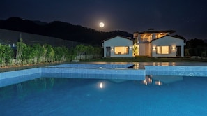A modern house with a swimming pool, a moon in the sky, and a fence with greenery.