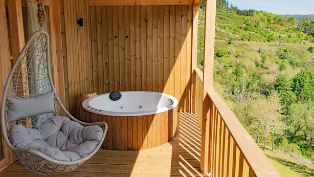 A wooden deck with a hot tub, a hanging chair, and a view of a green landscape.