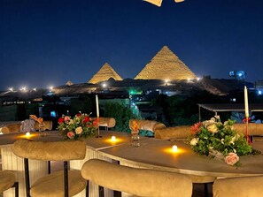A dining area with tables set for guests, featuring a view of two illuminated pyramids in the background. - Lavista Pyramid View (Giza)