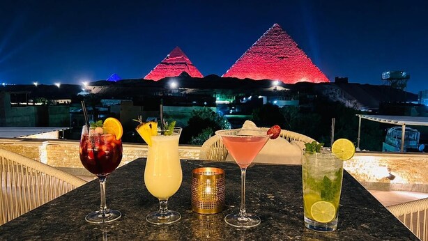 A table with five drinks, a golden candle, and two illuminated pyramids in the background. - Lavista Pyramid View (Giza)