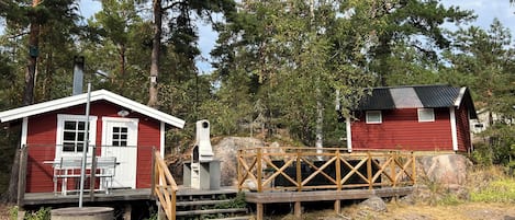 Una cabaña roja con un porche y una chimenea, un cobertizo pequeño y un puente de madera.