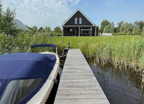 Eine Holzstege, der zu einem Haus mit dunklem Dach führt, umgeben von Wasser und Grün.