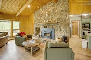 A cozy living room with a stone fireplace, wooden ceiling, and a view of the outdoors. - Mountain Views + Bbqs: Updated Cody Cabin (Cody)