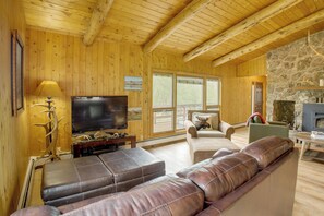 Interior de uma cabana de madeira com televisão, sofá de couro, lareira de pedra e janelas amplas.
