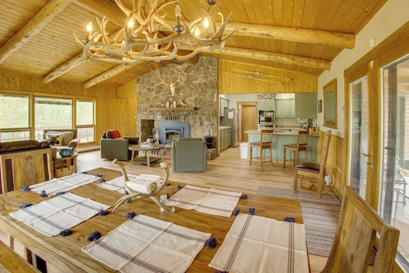 A spacious wooden interior with a stone fireplace, a dining area with a table set for four, and a kitchen visible through an open doorway. - Mountain Views + Bbqs: Updated Cody Cabin (Cody)
