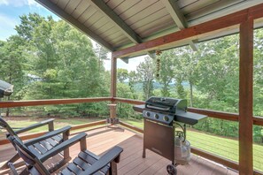 A covered outdoor deck with a grill, chairs, and a view of a forested area.