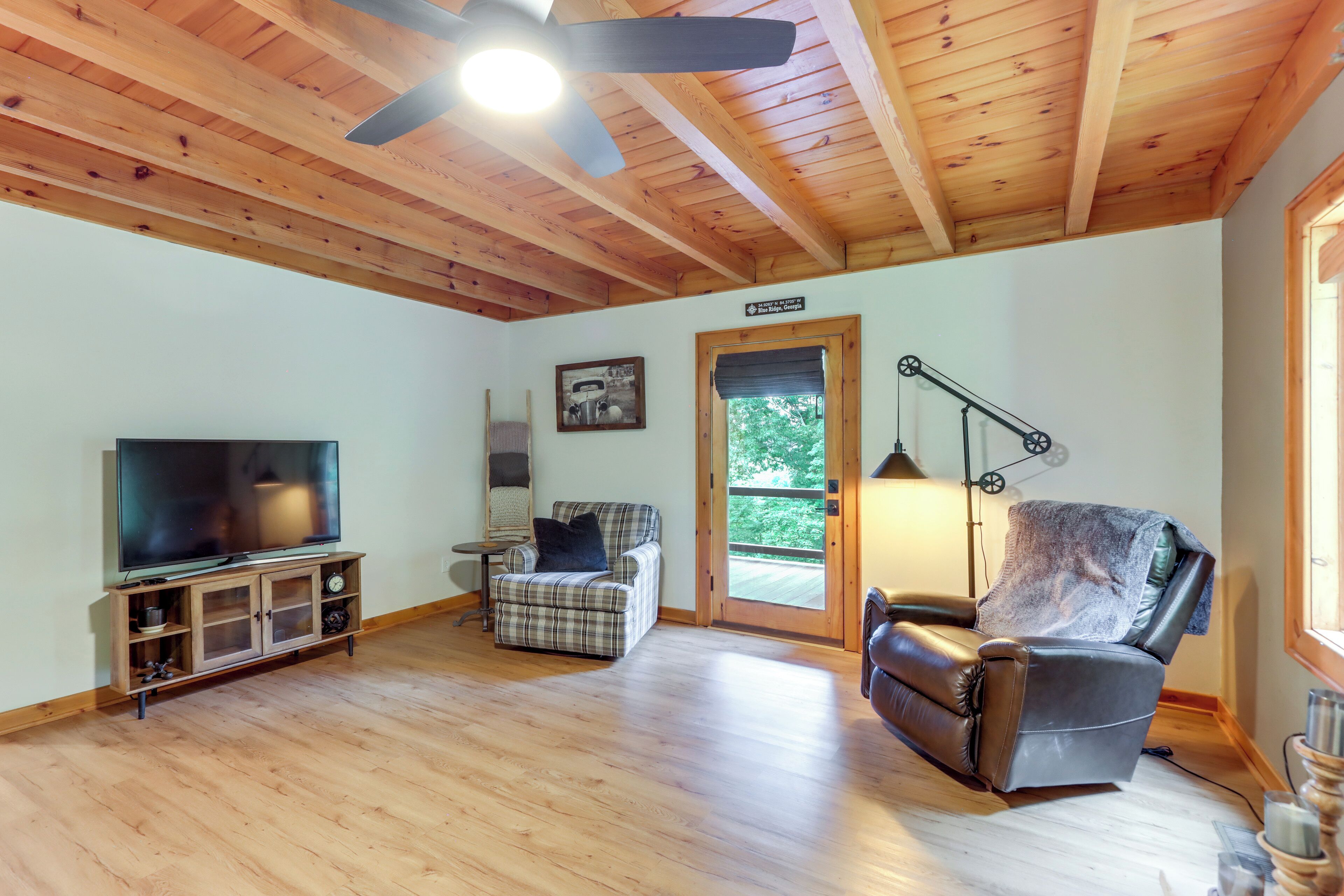 A living room with a wooden ceiling, a television, a plaid sofa, a leather recliner, and a door leading outside.