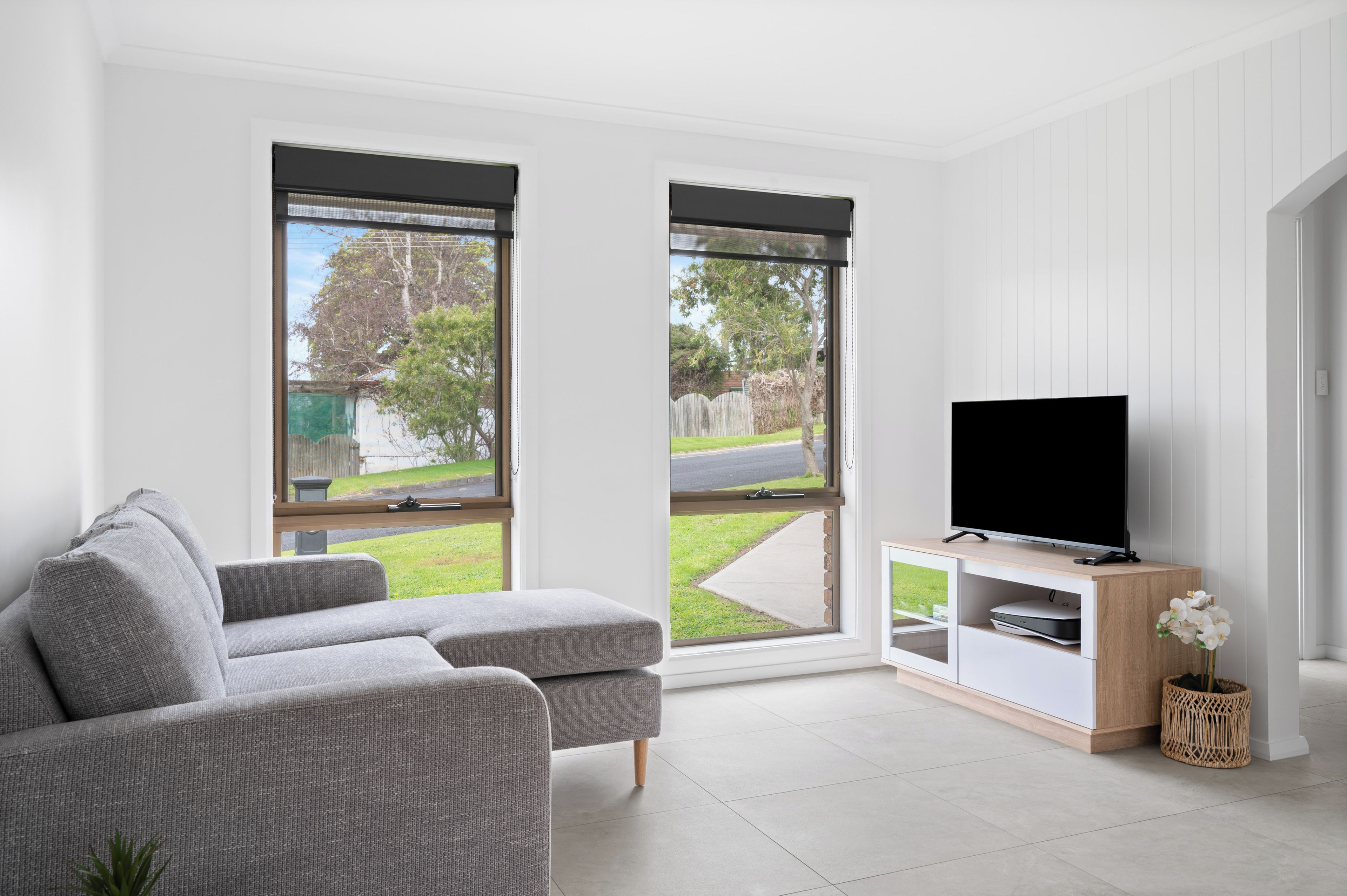A modern living room with a grey sofa, a flat-screen TV on a wooden stand, and large windows offering a view of a backyard.