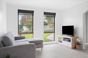 A modern living room with a grey sofa, a flat-screen TV on a wooden stand, and large windows offering a view of a backyard.