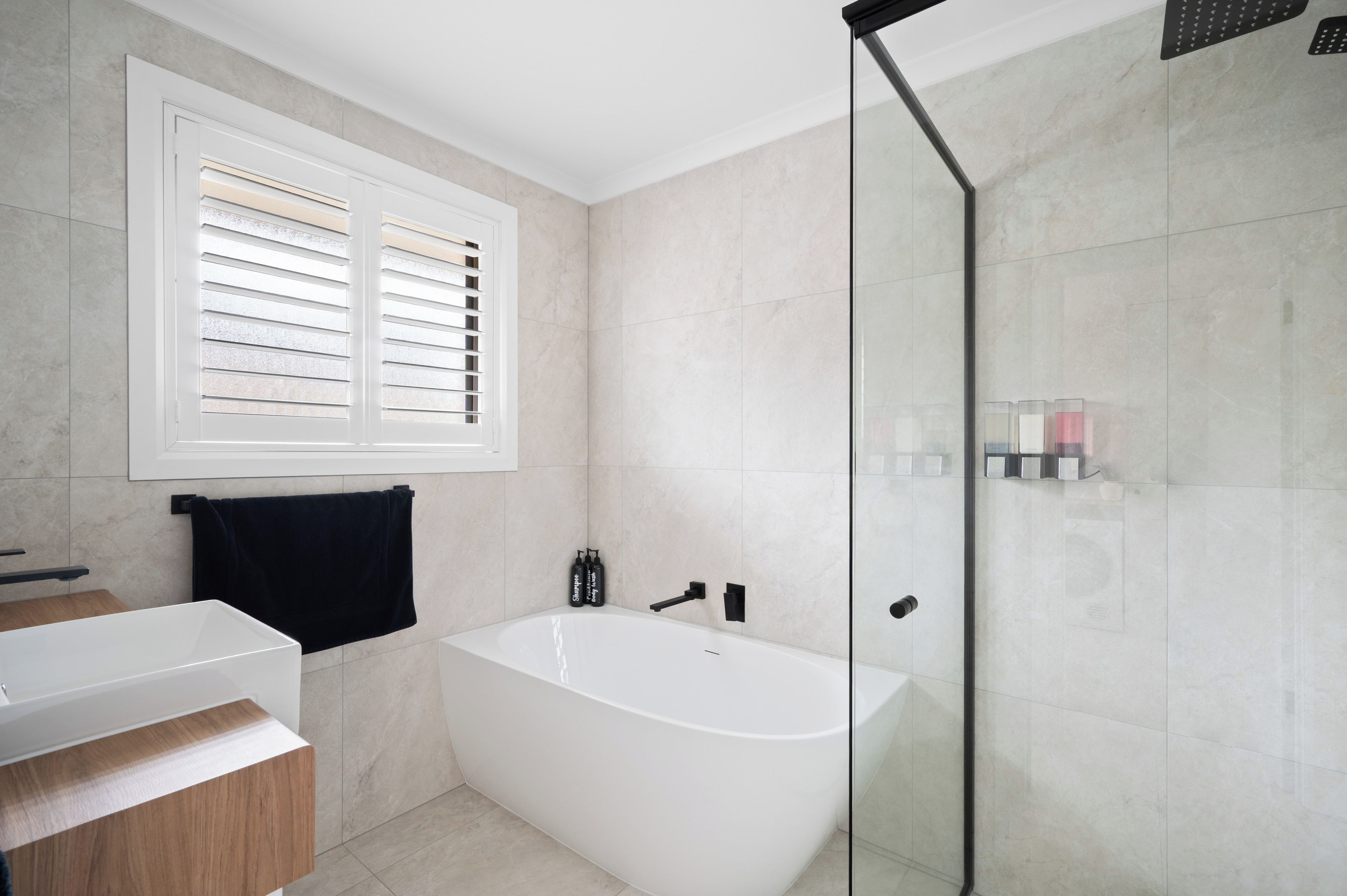 Ein modernes Badezimmer mit freistehender Badewanne, einer Glasdusche und einem Fenster mit Jalousien.