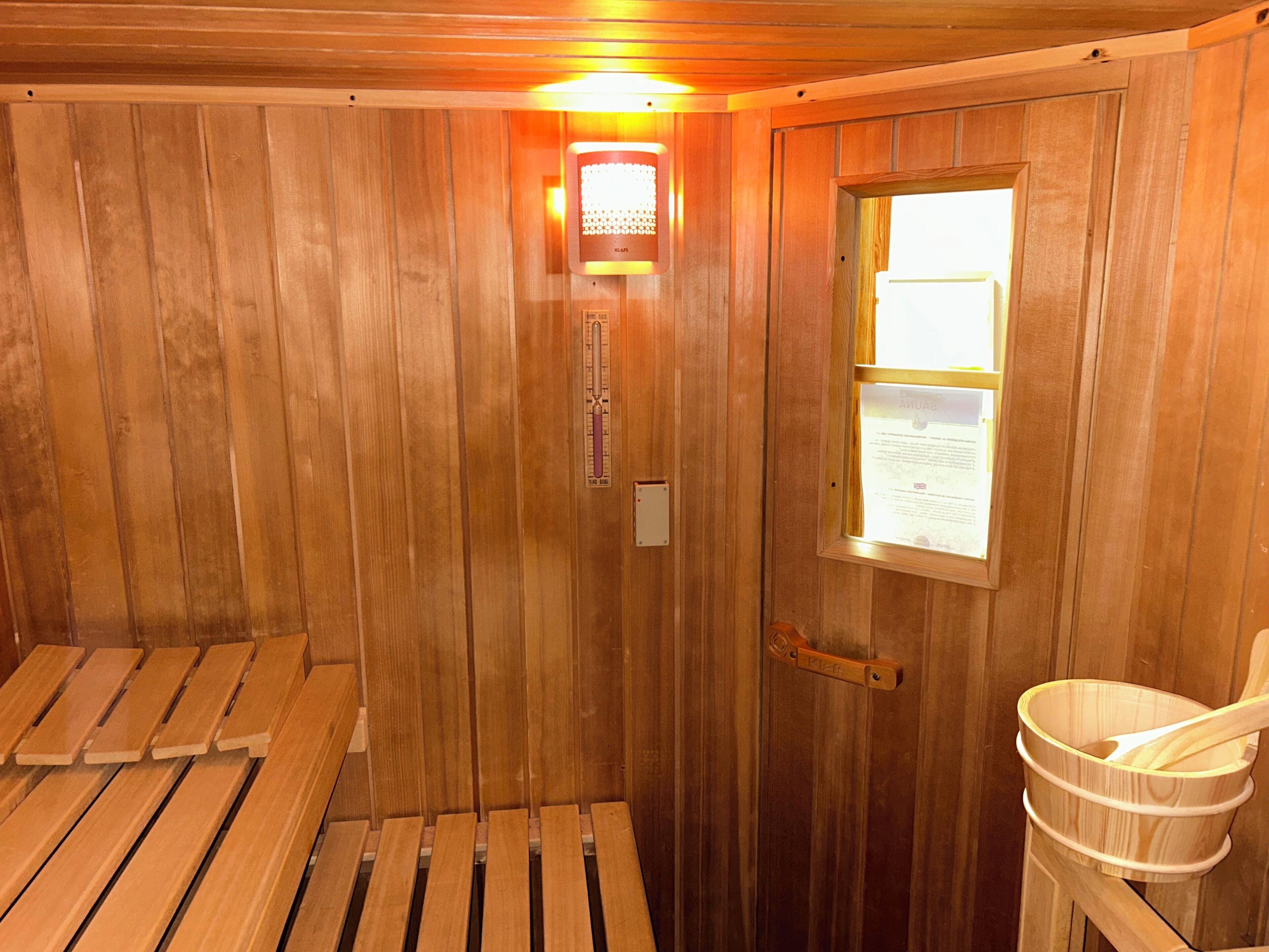 Een sauna met houten wanden, banken, een verwarming, een thermometer, een emmer en een deur met een raam.