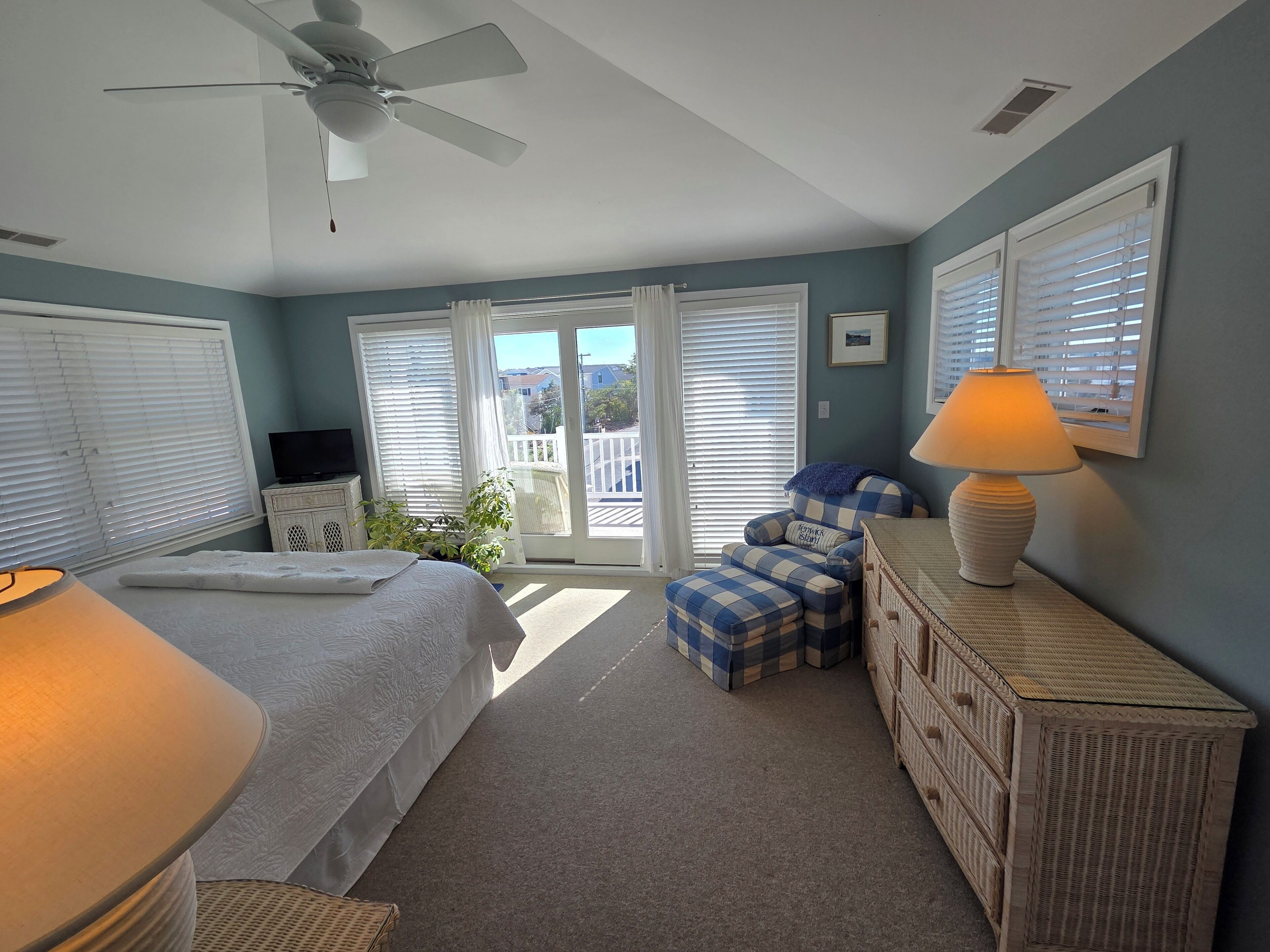 A bedroom with a bed, a chair, a dresser, a ceiling fan, and a balcony with blinds.