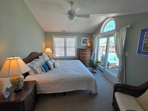 Ein Schlafzimmer mit einem Bett, Nachttischlampen, einem Schminktisch, einer Topfpflanze, einem Fenster mit Jalousien und einem Deckenventilator.