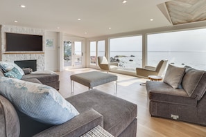 A spacious living room with a large window overlooking a beach, a fireplace, and a comfortable seating area. - Luxe 5,600-sq-ft Estate Waterfront + Pool (Westbrook)