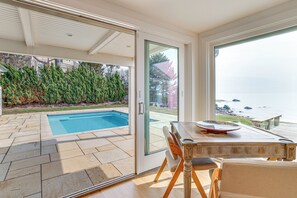 A poolside area with a table and chairs, a glass door leading outside, and a view of the ocean. - Luxe 5,600-sq-ft Estate Waterfront + Pool (Westbrook)