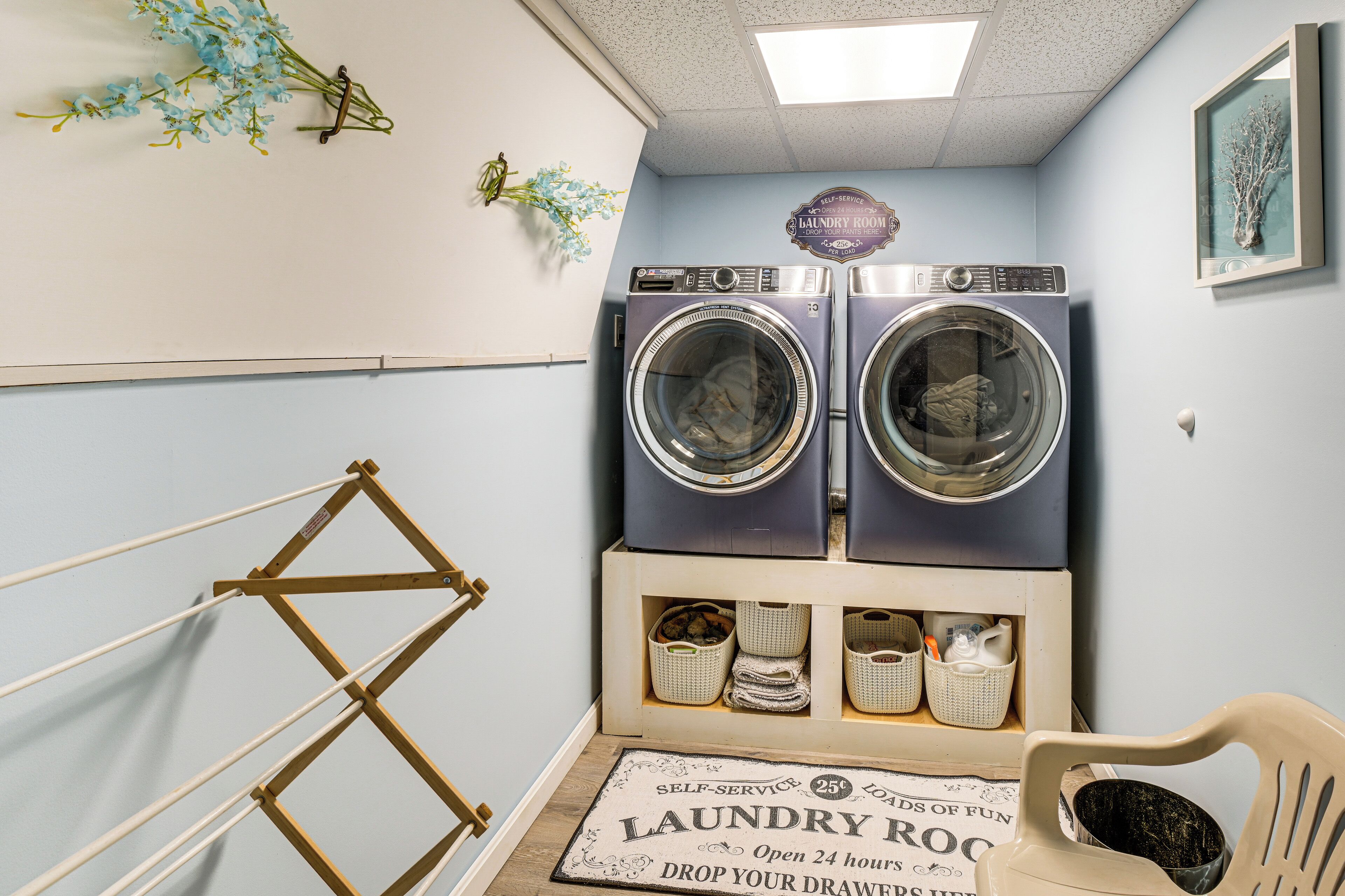 A laundry room with two front-loading washing machines, a drying rack, a wooden shelf with baskets, and a sign indicating self-service and 24-hour availability.