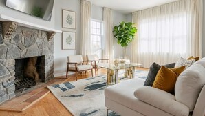 A living room with a stone fireplace, a white sofa, a dining table with chairs, and a large window with curtains.
