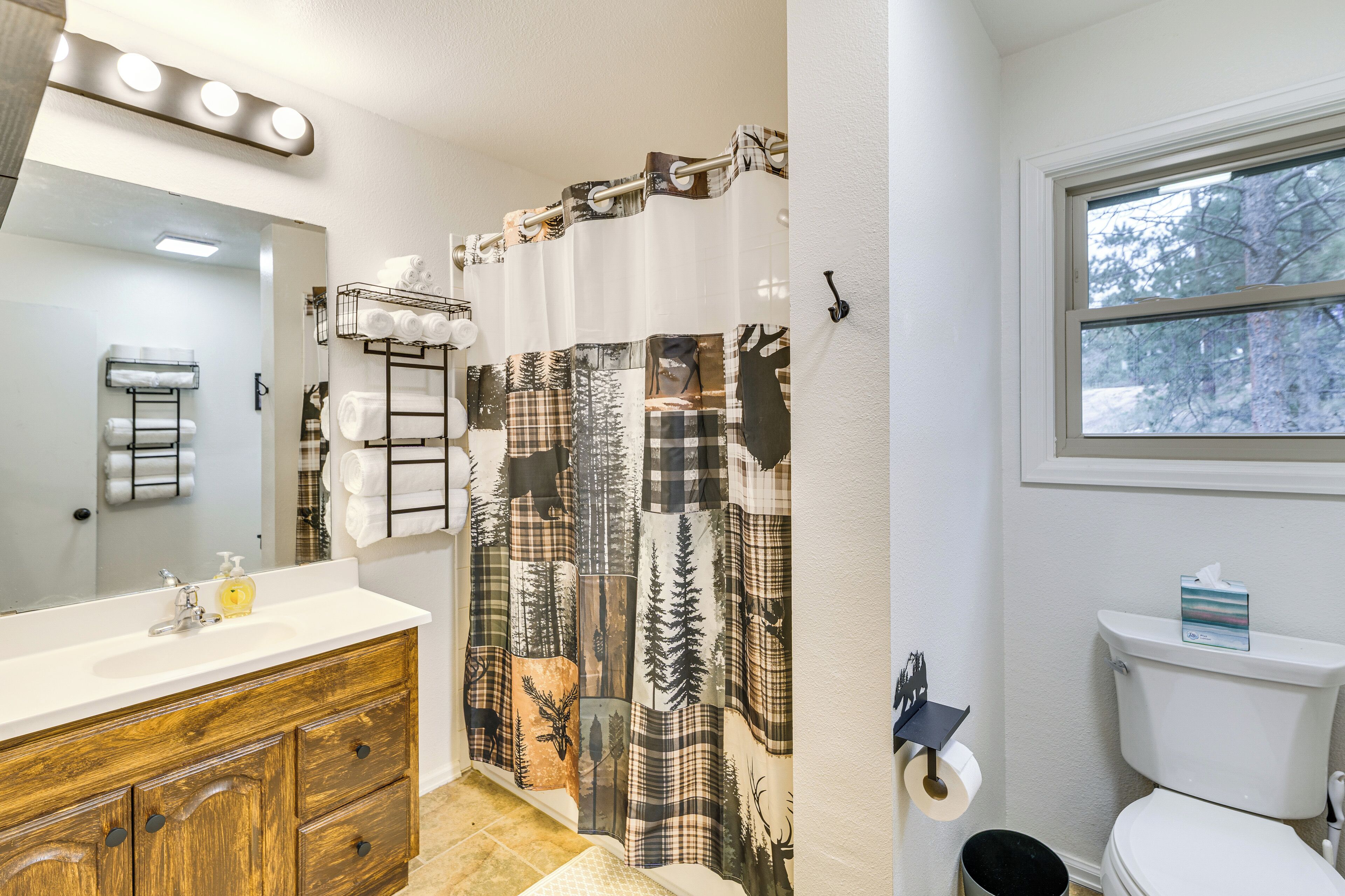 Un baño con tocador de madera, cortina de ducha con estampado de bosque, inodoro y ventana con vista a árboles.