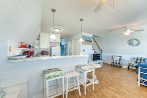 A kitchen with white cabinets, a bar area with stools, a dining area with a blue sofa, and a ceiling fan. - Walk to Waves! Surfside Beach House w/ Ocean View (Freeport)